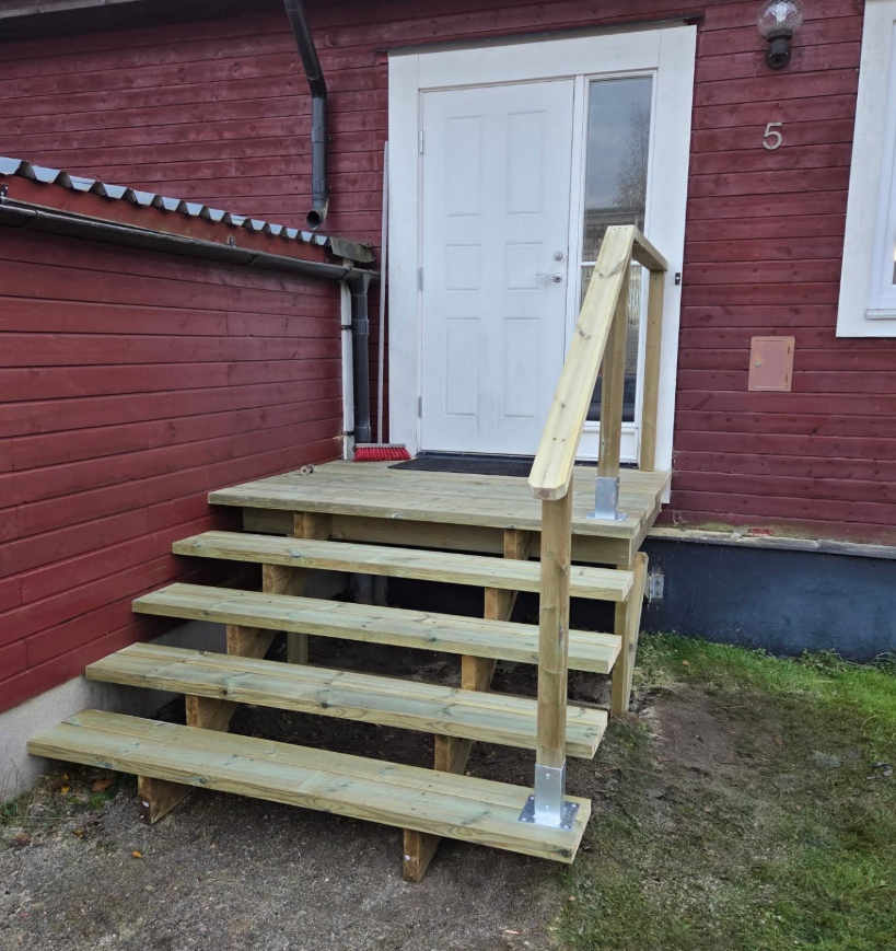 Ny entrétrappa i ljust tryckimpregnerat virke med räcke, mot röd träfasad.